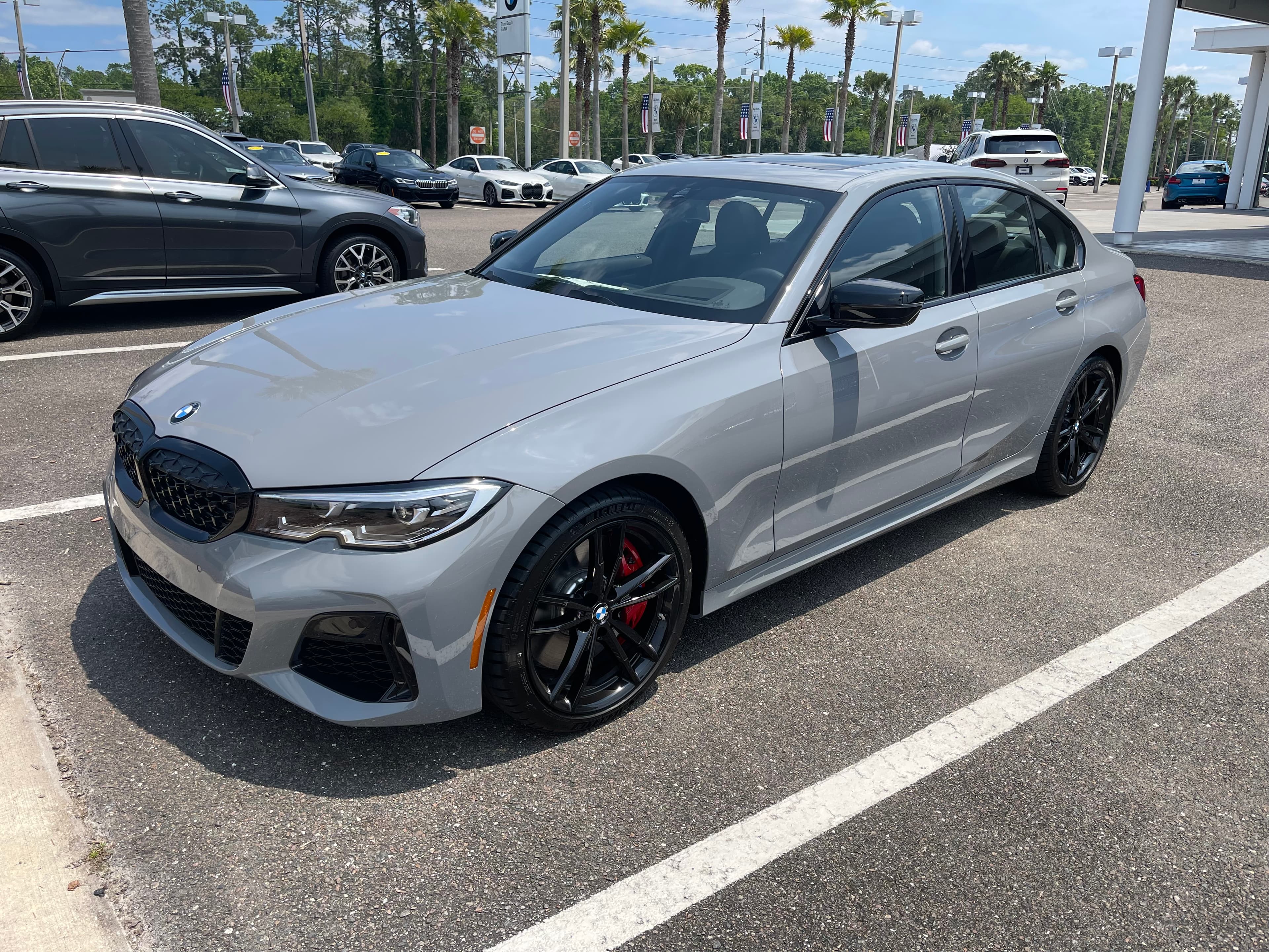 Grey BMW sedan with black wheels and red brake calipers at a sunny car dealership.
